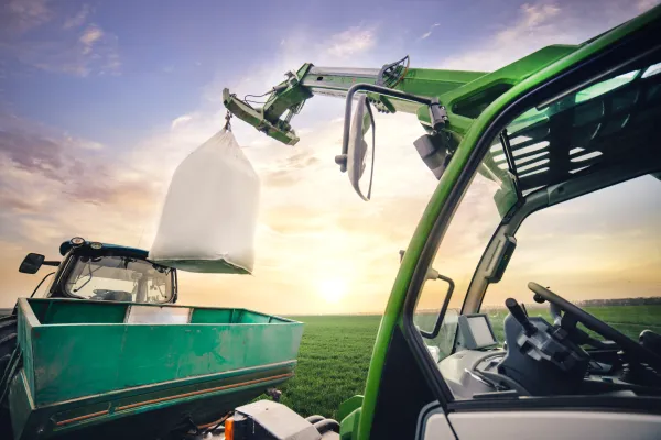Ein Kranarm lädt im Frühling auf einem Feld einen Sack Dünger auf den Anhänger eines Traktors